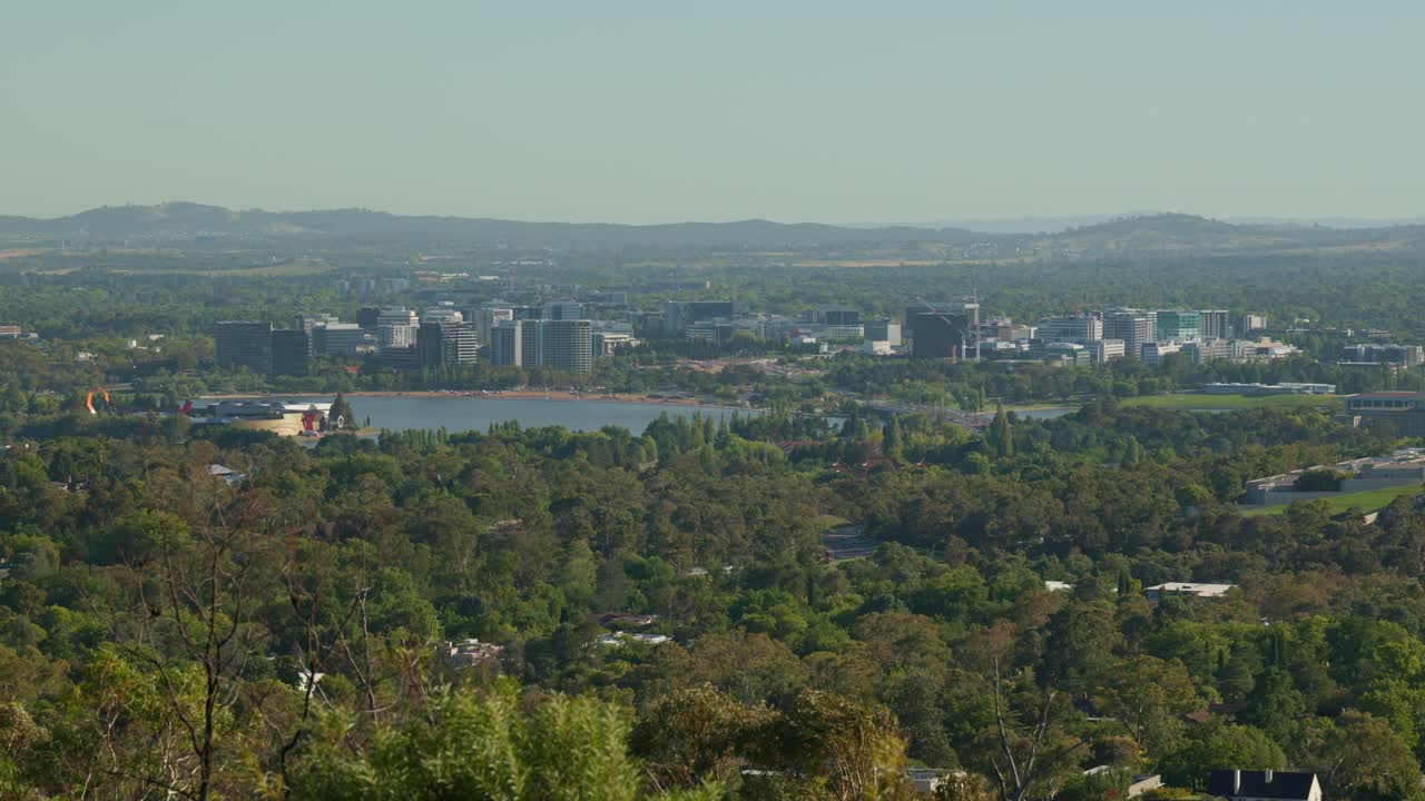 A slow zoom into Canberra’s city center, showcasing construction cranes, vehicles crossing the bridge over Lake Burley Griffin, and the capital’s urban growth in the morning light.