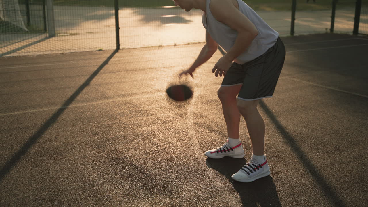 un jugador de baloncesto masculino rebotando y botando la pelota entre sus piernas en una cancha al aire libre al atardecer