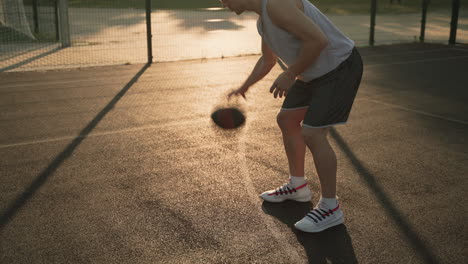 Ein-Männlicher-Basketballspieler,-Der-Bei-Sonnenuntergang-Auf-Einem-Außenplatz-Den-Ball-Zwischen-Seinen-Beinen-Hüpft-Und-Dribbelt