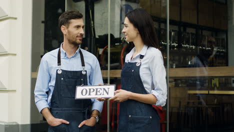 Cheerful-Young-Attractive-Waiter-And-Waitress-Standing-On-The-Street-At-The-Door-Of-The-Cafe,-Looking-At-Each-Other-And-Then-Smiling-To-The-Camera