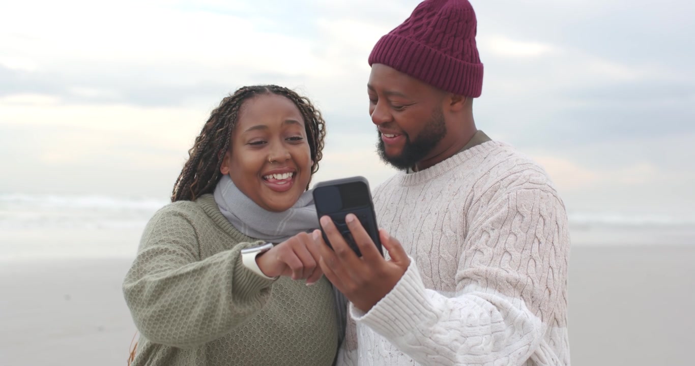 African American couple on beach in knits, beanie, man extending phone taking selfie, woman tapping