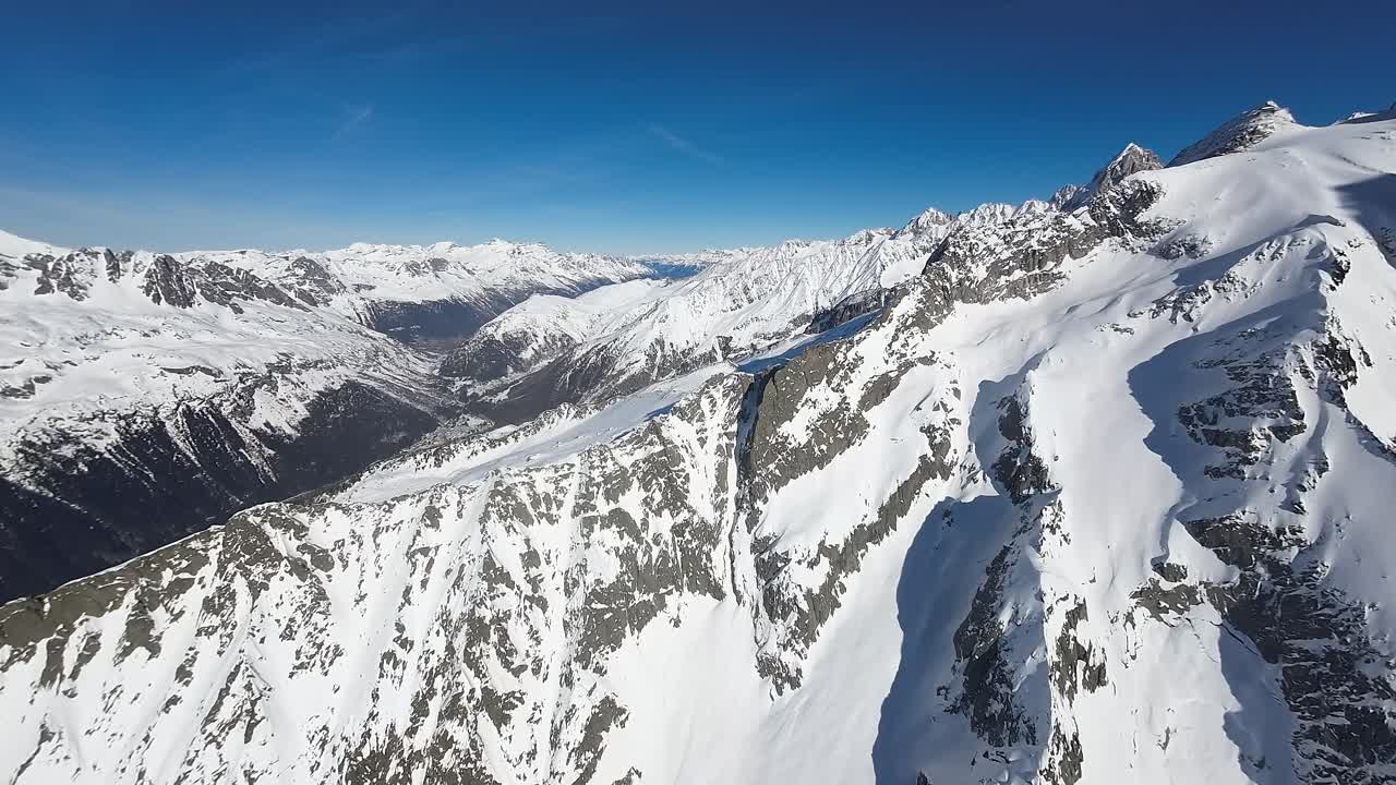 La vista aerea cattura la serena maestosità delle montagne innevate sotto un cielo azzurro e limpido, evidenziando la bellezza incontaminata di un paesaggio alpino invernale