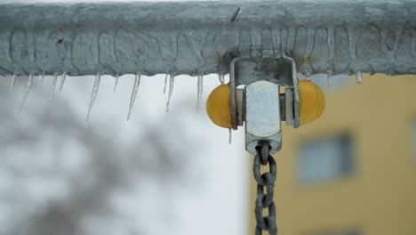 close-up of metal bar with icicles hanging, showcasing yellow plastic joint and chain detail, set against blurred snowy background and faint yellow building