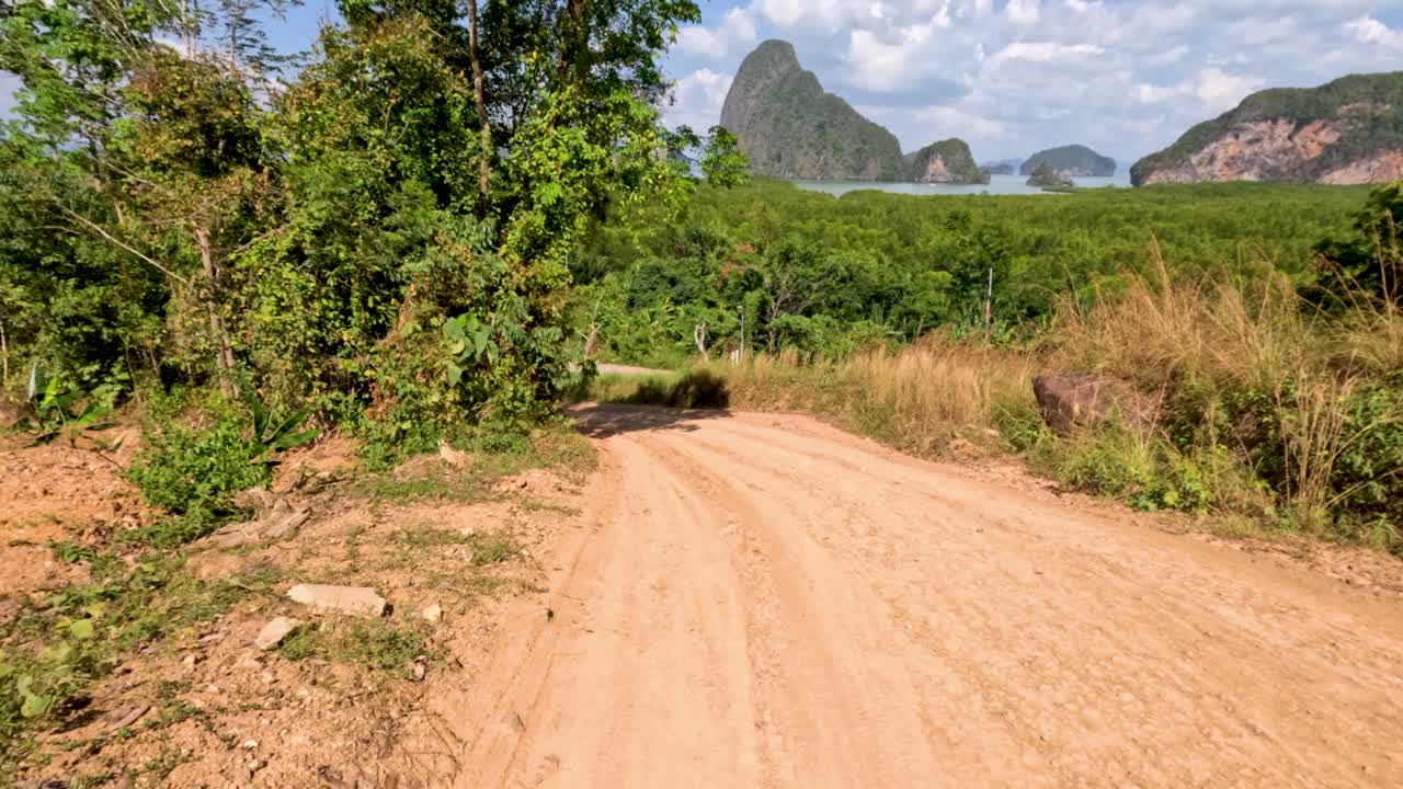 Vehicle travels along winding dirt road through sunlit tropical landscape, limestone cliffs visible ahead