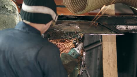 workman at a factory is grinding metal with circular saw on the bottom side of car construction