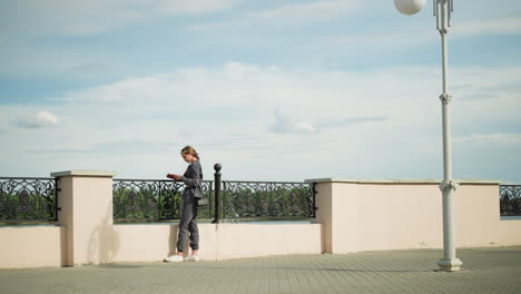 lady in grey clothing and black handbag stands close to a fence reading a book with focus and intent, trees are visible in the distance, and a lamp post is nearby