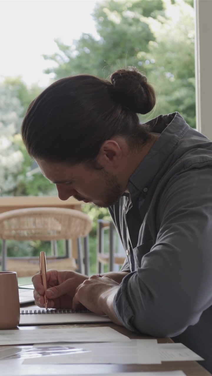 Vertikal video: Mann sitter ved bordet ved vinduet, gjennomgår og ferdigstiller notater i en spiralblokk