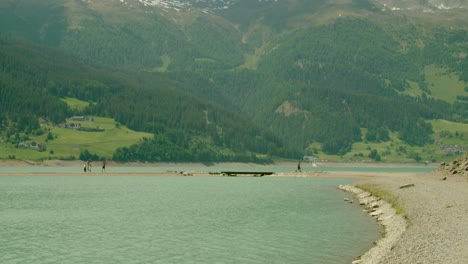 Full-shot,-People-walking-over-a-small-bridge-in-Reschensee-on-a-bright-sunny-day-in-Italy,-Scenic-view-of-mountain-landscape-in-the-background