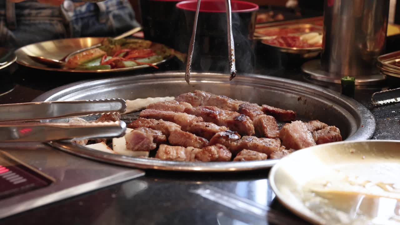 Pork belly pieces grilled on tabletop barbecue, tongs turning meat, warm indoor lighting, close-up view