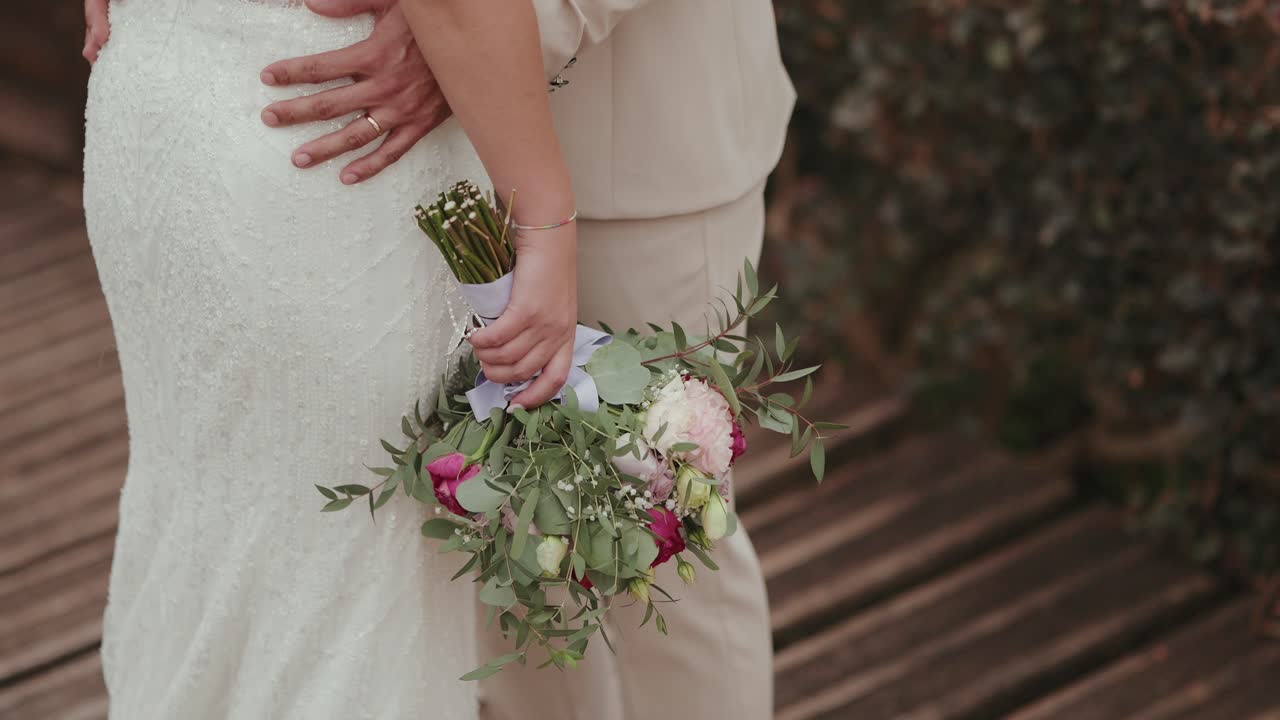Primo piano di una sposa in abito bianco che tiene in mano un bouquet di fiori mentre è in piedi accanto allo sposo che le cinge delicatamente la vita