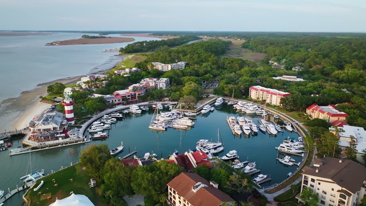 Aerial glide pullback over circular yacht marina passing lighthouse at tip of curved pier surrounded by calm harbor, Harbour Town Pier South Carolina USA