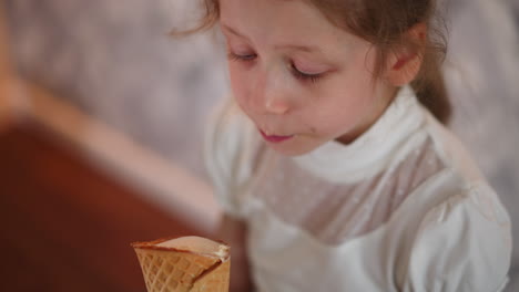 close up of white girl in white top enjoying ice cream cone, looking down thoughtfully while savoring her treat, standing near soft patterned wall