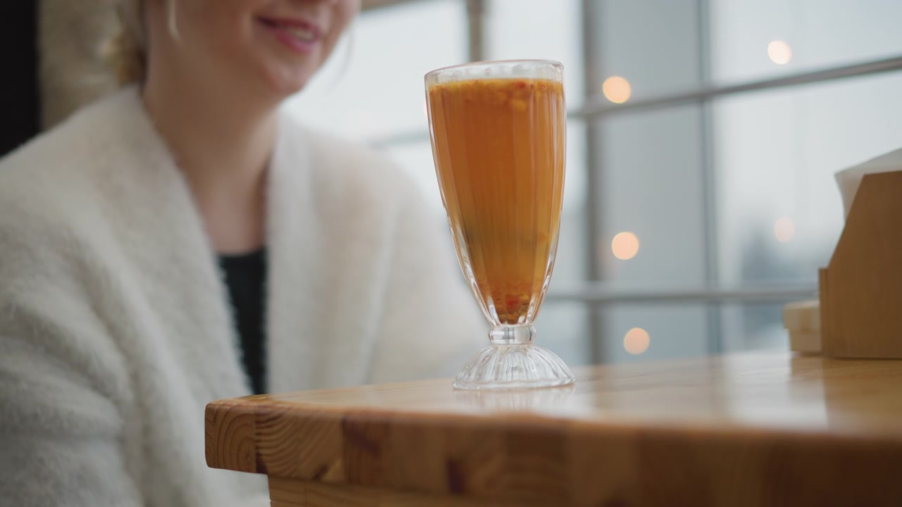Un calice ornato con una bevanda arancione poggia su un tavolo rustico di legno da caffè mentre una donna adulta sfocata siede dietro, creando una calda scena interna con condensa, un leggero increspatura, luce soffusa e un'atmosfera rilassata.