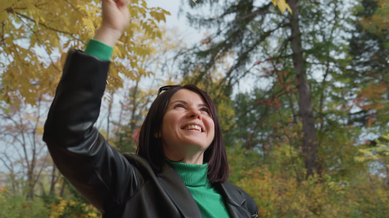 Mujer caucásica con chaqueta de cuero alcanzando hojas doradas de otoño en un parque iluminado por el sol, sonriendo ampliamente mientras mira hacia arriba, disfrutando del aire fresco otoñal, rodeada de árboles altos y follaje vibrante, natural