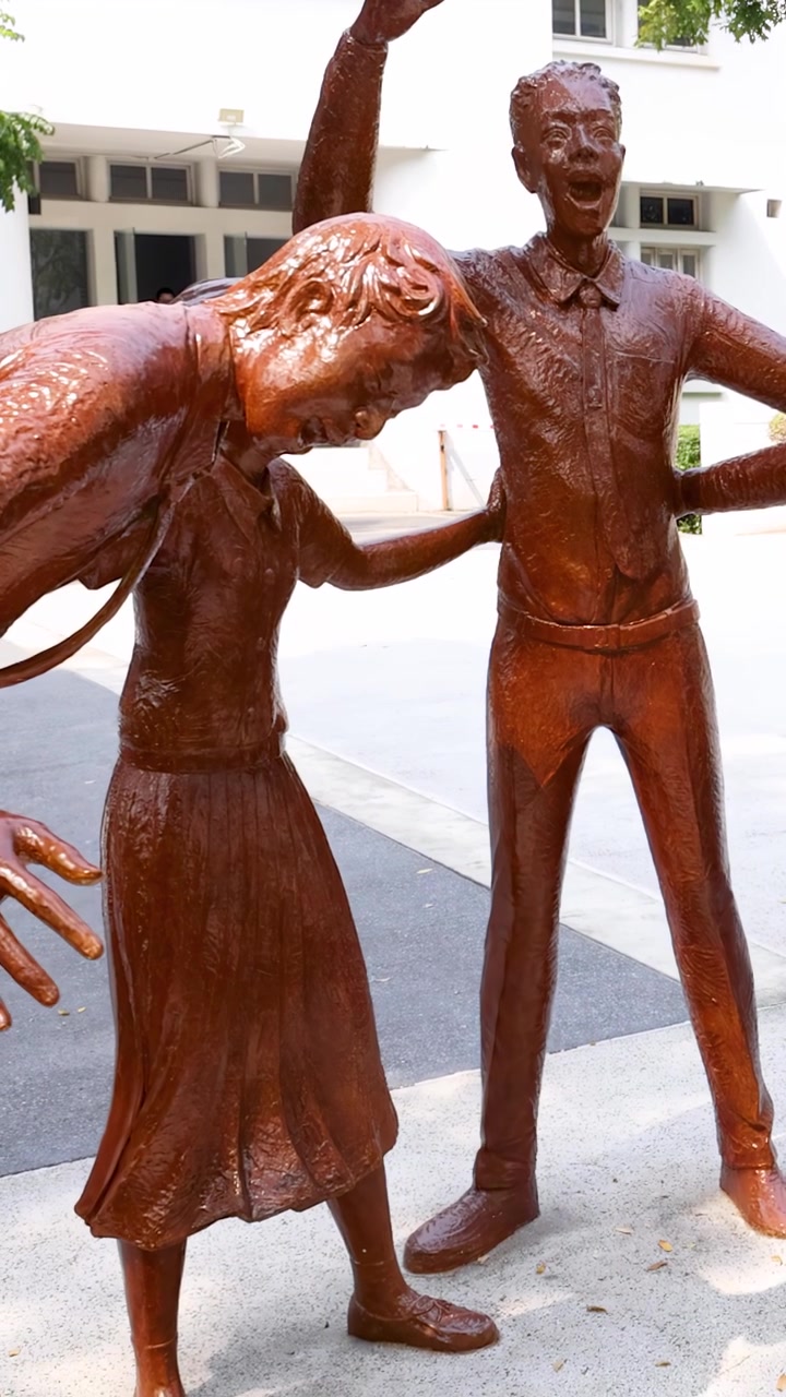 La telecamera zooma su statue di bronzo di studenti che ridono e giocano in un cortile esterno soleggiato.