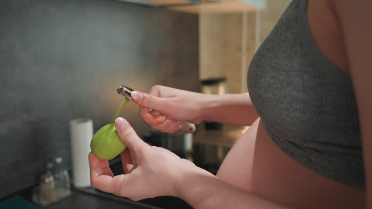 Mujer caucásica embarazada pelando una manzana verde en primer plano con el vientre visible, camiseta sin mangas gris en la encimera de la cocina, manos usando un pelador sobre la estufa, preparando un refrigerio saludable para el embarazo, táctil y concentrada.