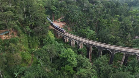 passenger train exits tunnel and onto nine arch bridge through the lush jungles of sri lanka. aerial.