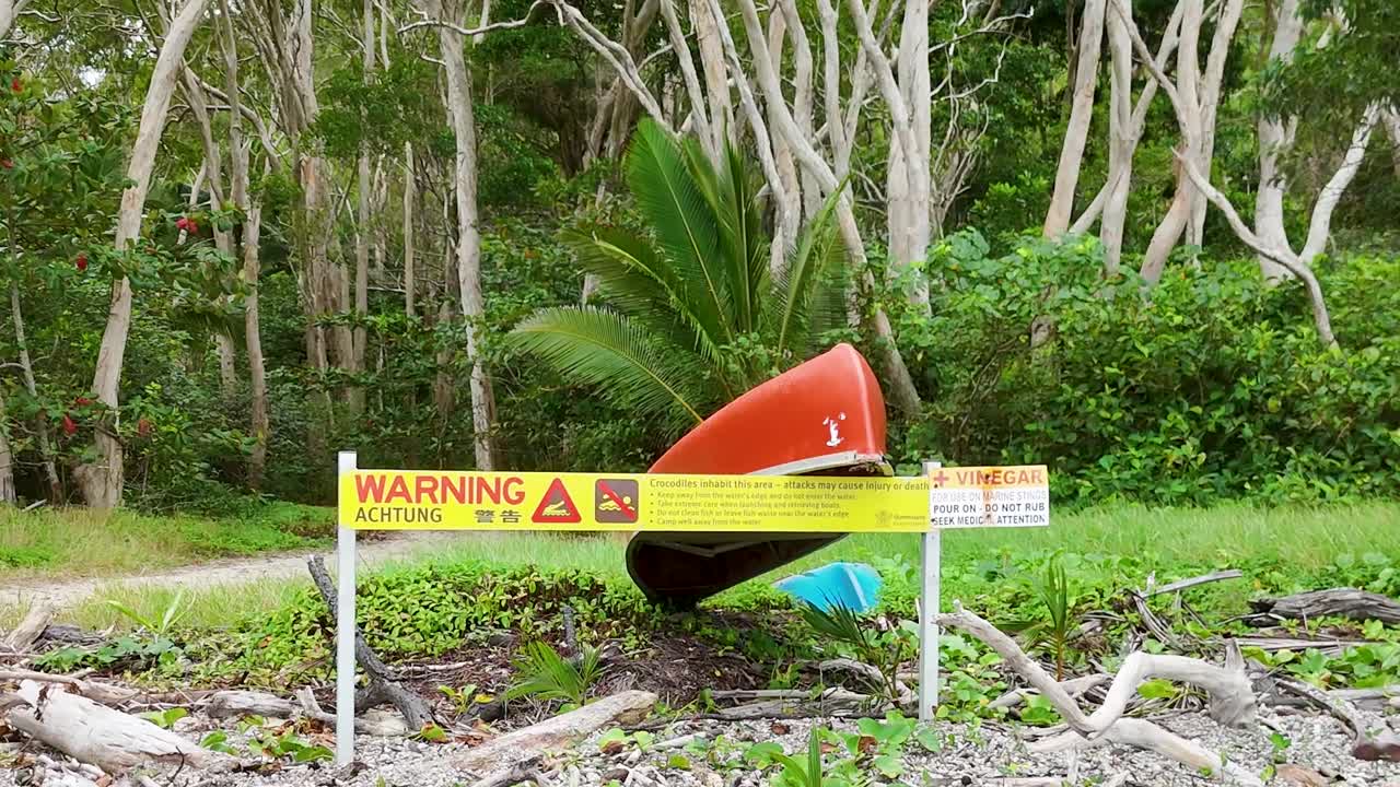 A yellow safety warning sign stands before a red boat hull in lush tropical greenery