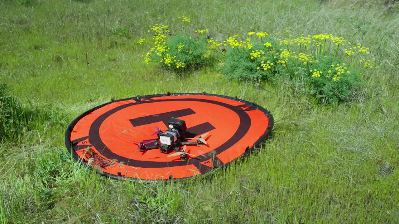 Un drone con visuale in prima persona (FPV) con una action camera GoPro montata che atterra su un eliporto arancione Hoodman mentre il vento soffia tra l'erba verde e i fiori gialli
