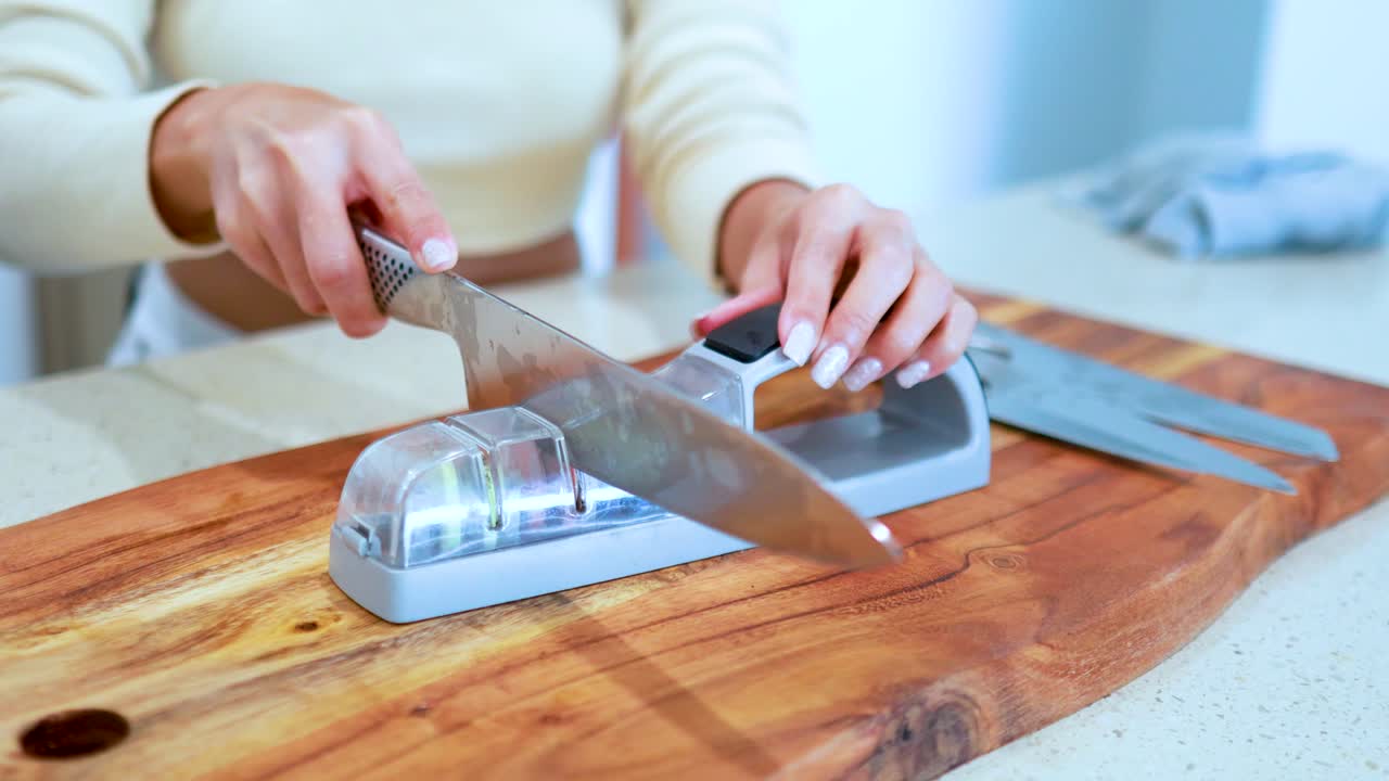 A person sharpens a large kitchen knife using a manual whetstone sharpener on a wooden cutting board in a brightly lit kitchen