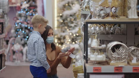 in a pandemic the coronavirus epidemic. a mother in a medical mask with her son at the mall choose home decorations for christmas. watch christmas tree toys. coronavirus epidemic