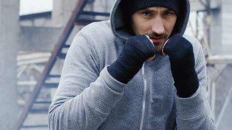 caucasian bearded caucasian man in grey hoodie and boxing to the camera outdoors an abandoned factory on a cloudy morning