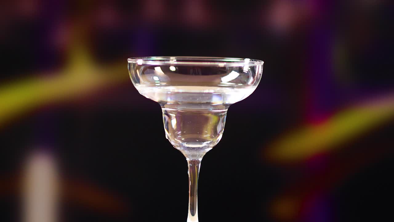 una mano coloca un giro de limón en un vaso de martini vacío contra un fondo borroso y colorido