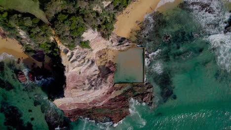 top-down drone view slowly descending over aslings beach rock pool, revealing colorful cliffs, twin beaches, and the lush grassy headland above. captured in eden, nsw