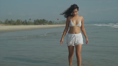 Slow-motion-clip-of-south-asian-woman-walking-through-shallow-sea-water-on-the-beach-shoreline,-towards-the-camera,-wearing-white-bikini