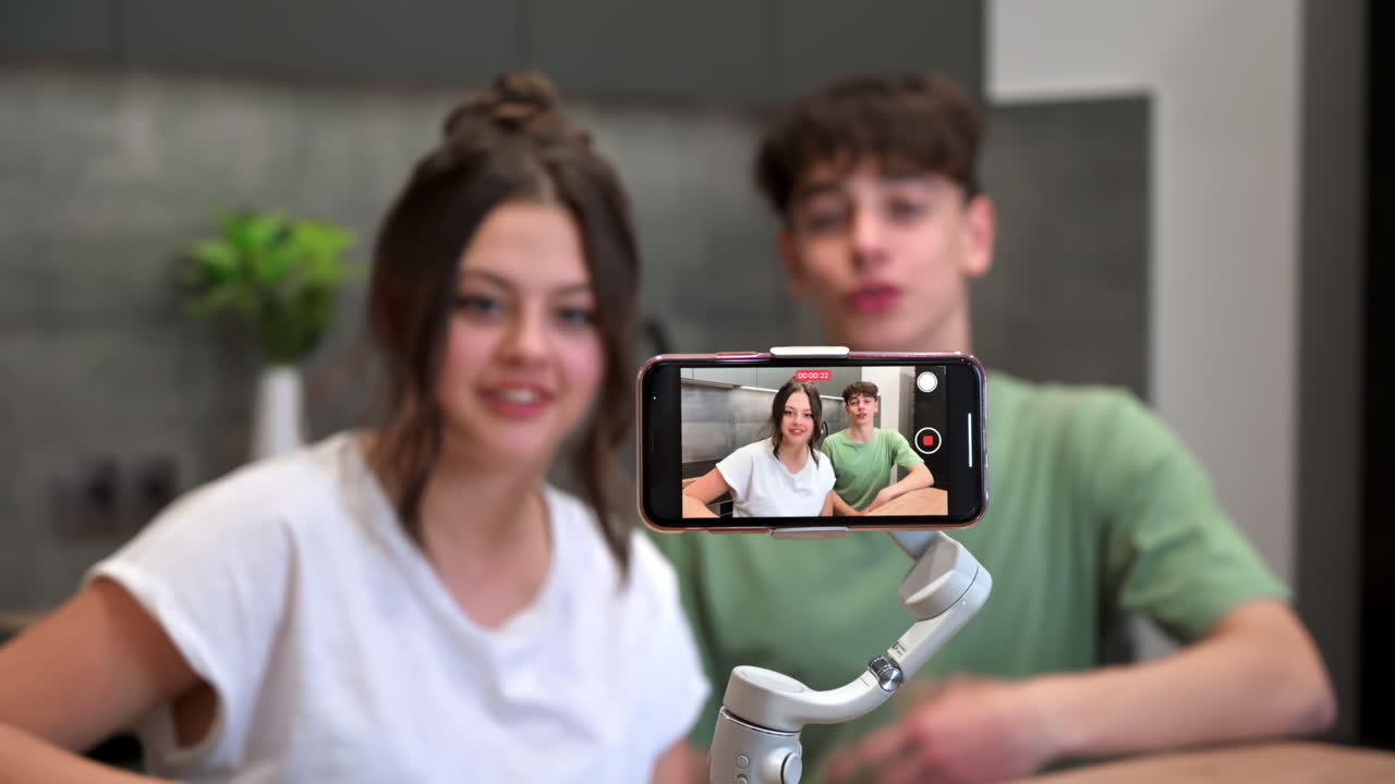 Giovane uomo e donna sorridenti, blogger e influencer, che parlano e si filmano con lo smartphone su uno stabilizzatore in una cucina