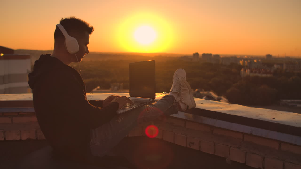 программист с ноутбуком в наушниках сидит на крыше и пишет код на закате и слушает музыку. удаленная работа фрилансер. свобода работы. набирать на клавиатуре на закате с видом на город.