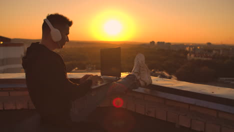 a programmer with a laptop in headphones sits on the roof and writes code at sunset and listens to music. remote work freelancer. freedom to work. typing on a keyboard at sunset with a view of the city.