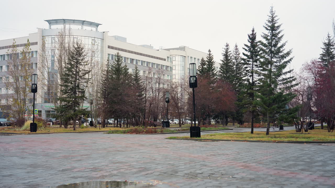 Empty paved urban park area surrounded by evergreen and bare trees near modern building on cold overcast day, light snowfall gently falling and reflections in shallow puddles on ground