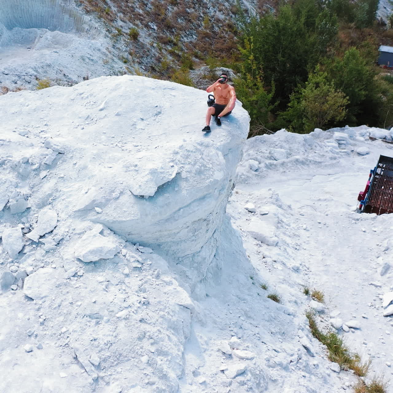 Brutal strong bodybuilder posing outdoor. Photoshoot in a quarry. Outdoor sports concept. Aerial view. Handsome strongman sits on a peak of a quarry. White landscape.