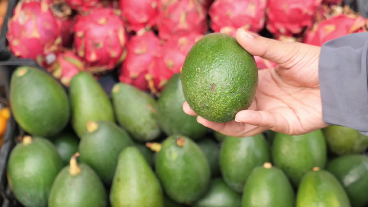 persona sosteniendo un aguacate frente a una exhibición de aguacates y frutas del dragón