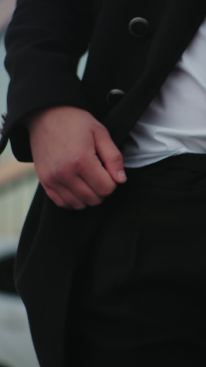 검은 코트를 입은 남자가 주머니에 손을 넣고, 주차된 자동차와 건물을 특징으로하는 배경, 도시 환경에서 자신감 있고 세련된 개인, 정교함과 전문성을 발휘합니다.