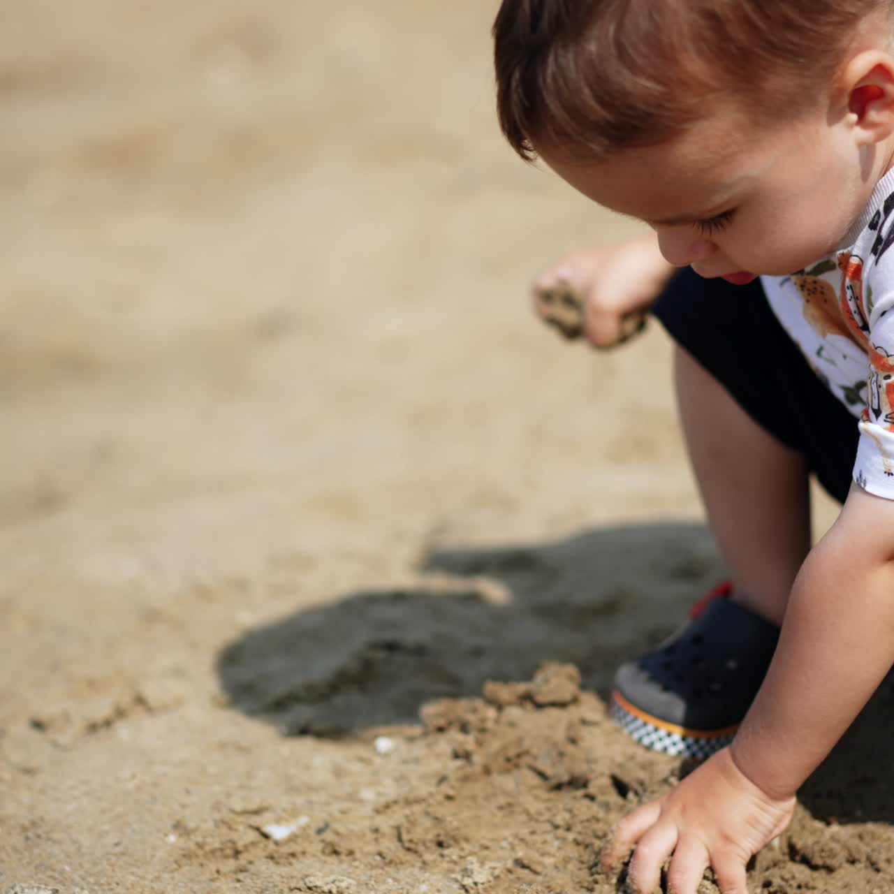 Beautiful Caucasian toddler squatted on the shore. Baby picks wet sand throwing it away again