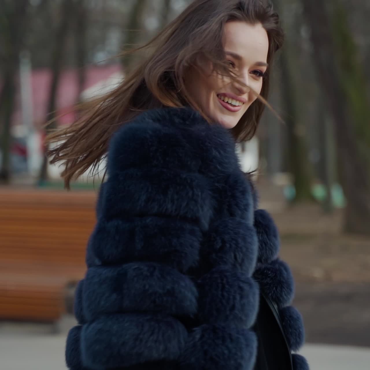 Glamour model posing on camera outdoors. Portrait of beautiful girl in a black fur coat smiling on the park background. Motion camera around.