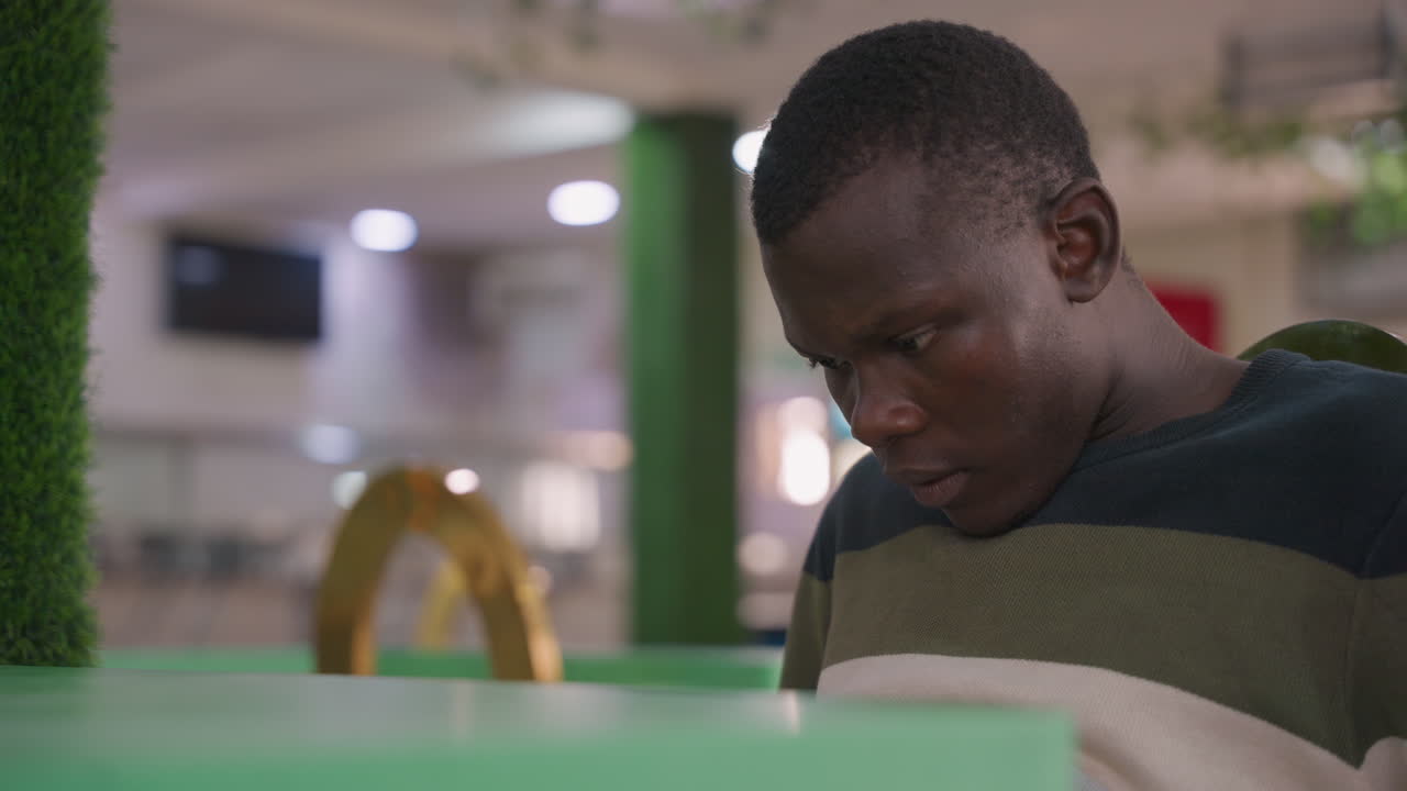 Man touching ear at mall table, practicing words under breath, focused expression with striped sweater and green table foreground, soft lights and quiet crowd creating pensive rehearsal atmosphere