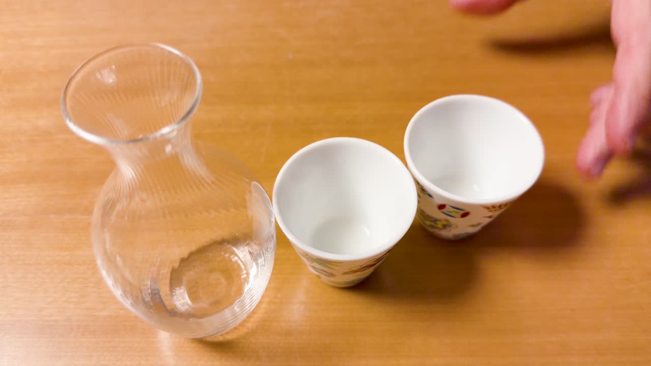 Una mano posiziona una tazza ochoko in ceramica accanto a una caraffa di sake in vetro su legno