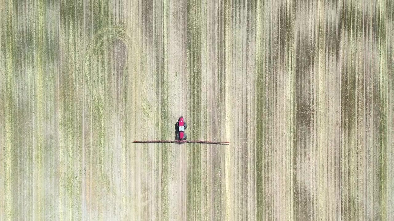 Aerial top-down view of a farm sprayer moving across patterned crop field rows, creating precise agricultural coverage. The geometric lines of the field highlight modern farming efficiency, precision
