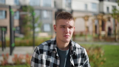 ill man in checkered shirt takes pill under bright sunlight, squinting eyes slightly closed while standing near blurred residential buildings with green outdoor surroundings in urban setting