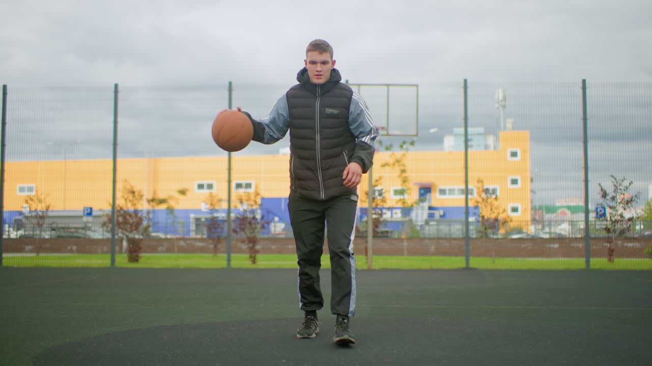 Un giovane uomo palleggia con un pallone da basket sul campo sotto un cielo nuvoloso, parco giochi urbano recintato con edificio giallo sullo sfondo, indossa un piumino e scarpe da ginnastica, allenamento individuale con concentrazione