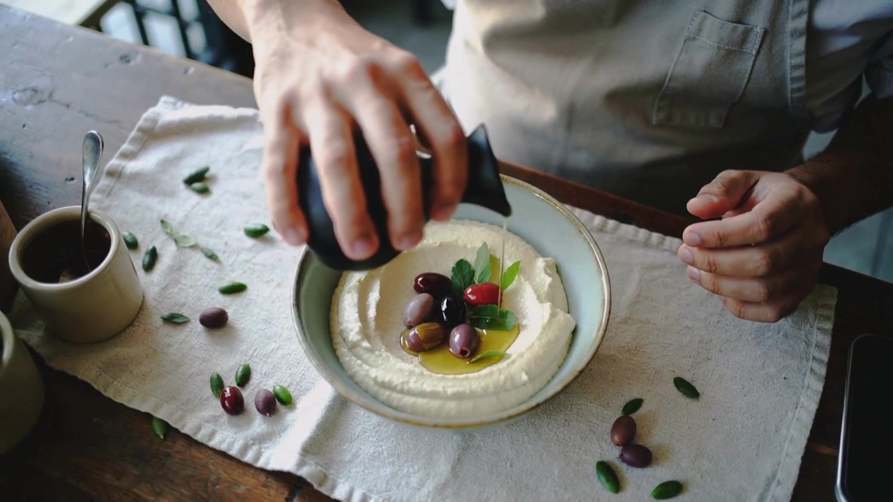 Chef con grembiule che dispone olive, afferra un'oliera nera e versa l'olio in una ciotola su un tavolo di legno, completando la preparazione