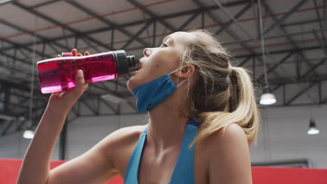 Kaukasische-Frau-Mit-Abgesenkter-Gesichtsmaske-Trinkt-Wasser-Im-Fitnessstudio