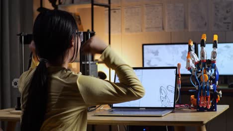 vista trasera de una adolescente asiática con impresión 3d celebrando el éxito en el diseño de una mano cyborg en una computadora portátil en casa