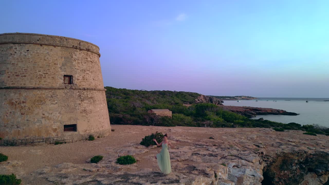 Woman performing yoga poses at sunset near Tower and Es Vedra Island in Ibiza, Spain. Tremendous aerial view flight ascending drone