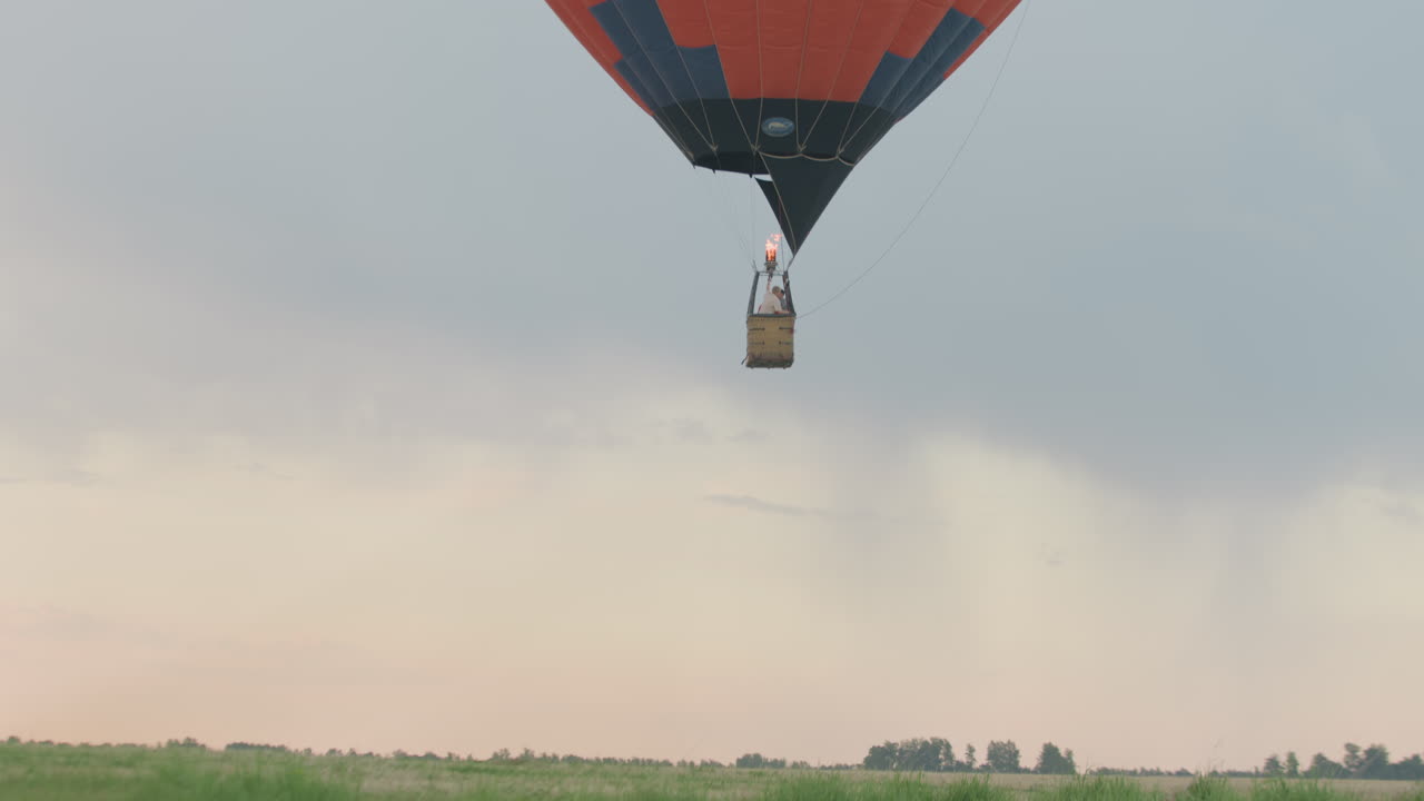 People enjoying sunrise flight in vibrant hot air balloon, passengers peering over wicker basket edge, pilot adjusting burner flame, floating above green open field under pastel dawn sky