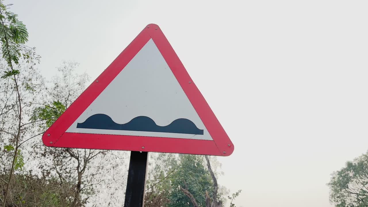 Camera orbiting around a triangular road humps ahead warning sign on a roadside pole, red border and wave symbol visible against pale sky and trees in daylight