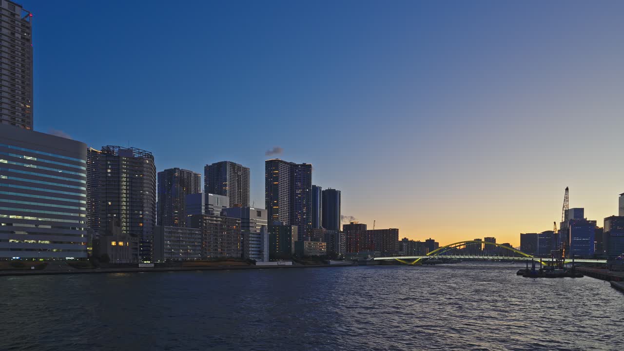 Uma vista noturna serena da orla de Tóquio ao crepúsculo. O rio calmo reflete a luz que se esvai enquanto a cidade transita para a noite. Arranha-céus modernos alinham o horizonte, e uma ponte em arco distinta.